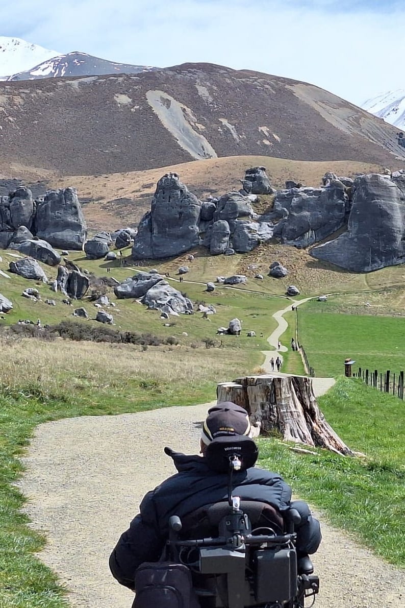 A man in an electric wheelchair on a path with a mountain range and large boulders ahead