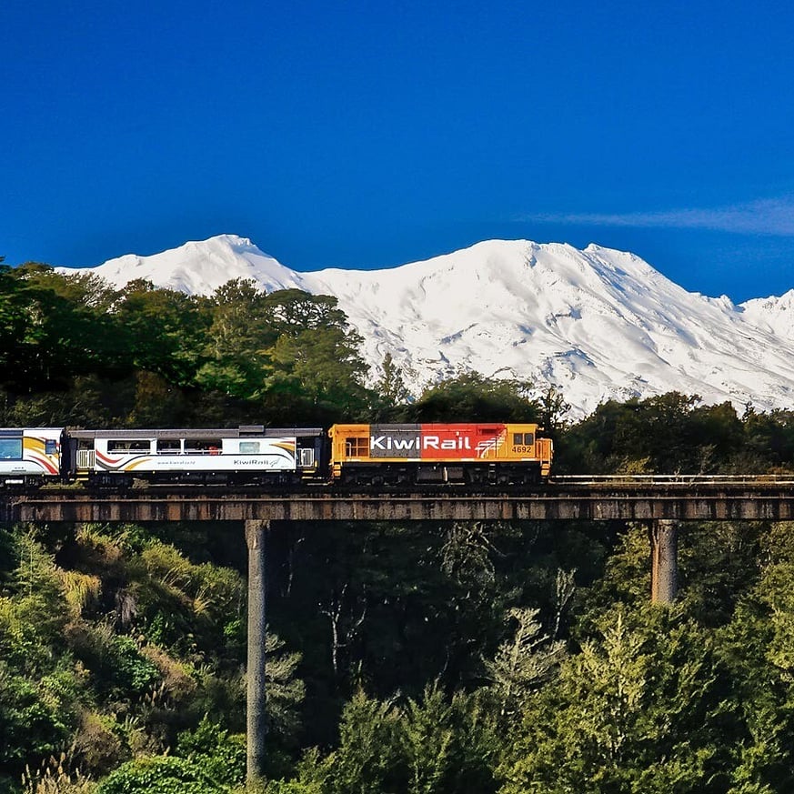 Northern Explorer train on a scenic journey with snow-covered mountains, possibly Mount Ruapehu, in the background