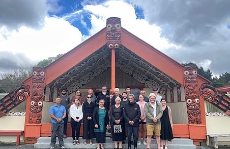 Group of adults standing outside carved Māori wharenui with red and black detailing and traditional patterns under cloudy sky