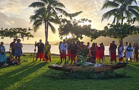 Group gathered outdoors by water at sunset with palm trees and cultural ceremony around decorated canoe on grass