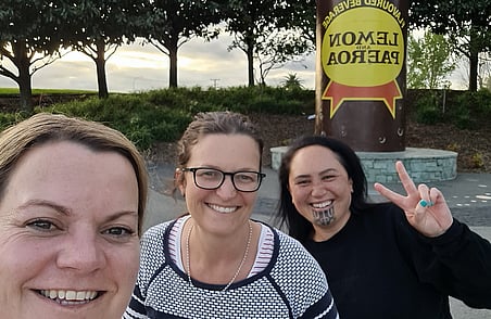 Three women smiling for selfie outdoors near Lemon and Paeroa sign with trees and evening sky in background