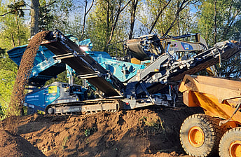 Powerscreen Warrior 1200 screen machine working in a forest to process soil into grades and being loaded by a moxy tip truck