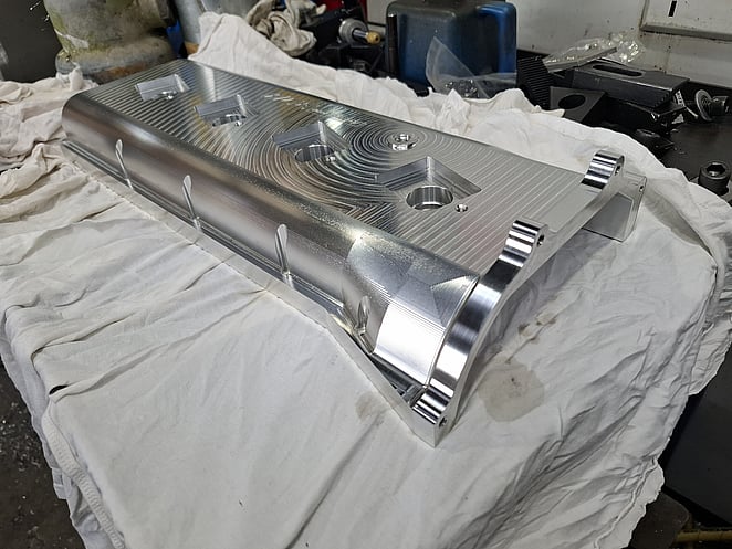 Precision-machined aluminium component with multiple cutouts and curved edges, resting on a white cloth.