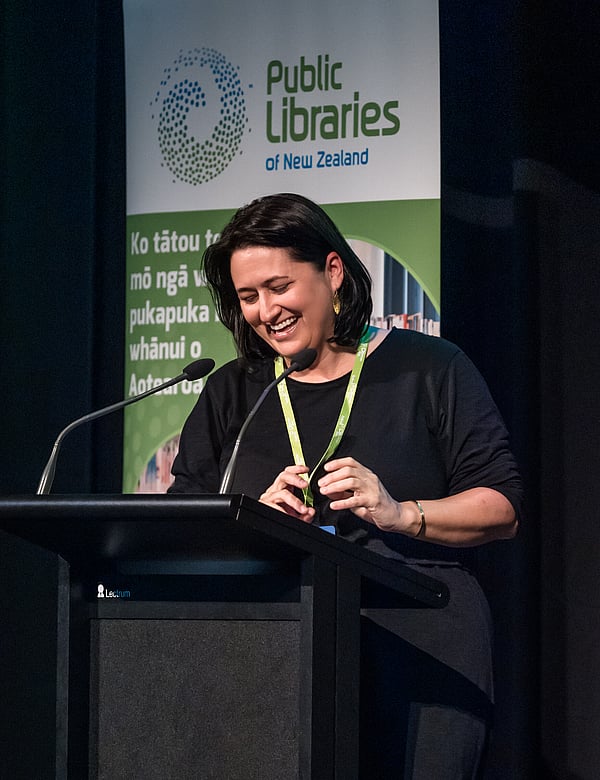 Event presenter laughing at podium with event banner in background. Public Libraries NZ Forum in Rotorua, NZ