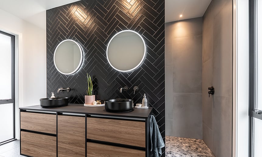 Modern black bathroom featuring a floating wall, black herringbone tile, with circular lit mirrors, and a walk-in pebble shower.
