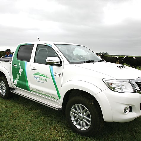 White ute with green and blue decals, map of New Zealand and Open Country logo on side door, in a paddock with cows behind.
