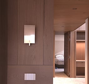 Tiled floor hallway with warm timber walls and ceiling, wall lights and switches in focus, opening to a bedroom with a detailed timber cross-pattern door in an Ocean Beach Road coastal home built by MB Construction.