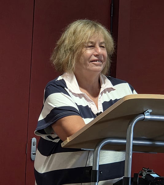 Woman with blonde hard standing at a lectern. She is smiling.