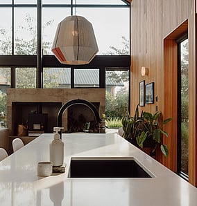 Kitchen in an architectural rural home near Matamata with island bench flowing through to the living room, concrete fireplace and window seat framing the rural outlook, built by MB Construction, residential builders for Matamata and the Bay of Plenty.