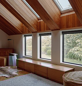 Light-filled architectural rural home bedroom in Matamata with raked timber ceiling, twin skylights and a built-in window seat framing countryside views, by MB Construction.