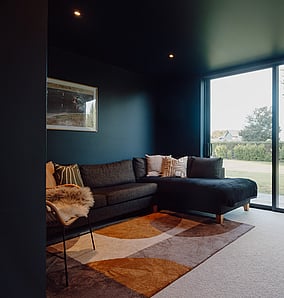 Media room in an architectural rural home in Matamata with dark walls, built-in cabinetry and a large window looking to the garden, designed and built by MB Construction.