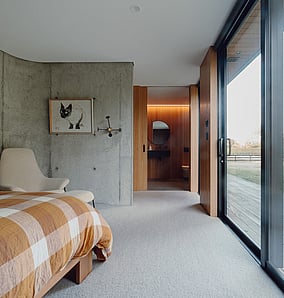 Main bedroom and ensuite threshold in an architectural rural home in Matamata, showing concrete feature wall, timber flooring and open connection to the ensuite, crafted by MB Construction.