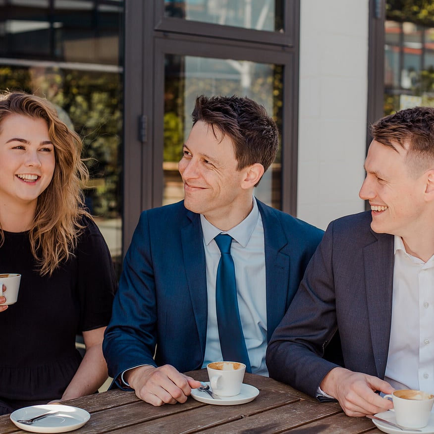 Three professionally dressed people sitting at an outdoor caf&eacute; table, smiling and chatting while holding cups of coffee