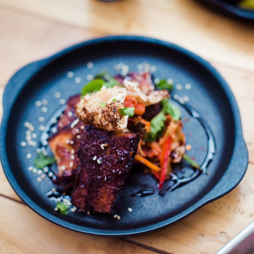 Black plate with glazed pork belly, crispy topping, and fresh salad, garnished with sesame seeds, on a wooden table with a glass of beer