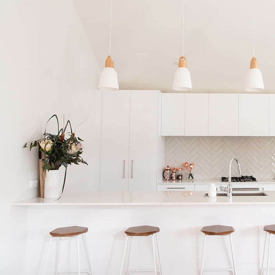 Modern white kitchen with a large island, wooden bar stools, pendant lights, and a vase of fresh flowers on the counter