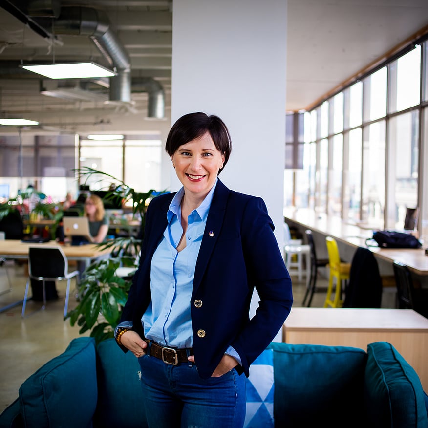 Smiling woman in a navy blazer and blue shirt standing in a modern coworking space with large windows and people working in the background
