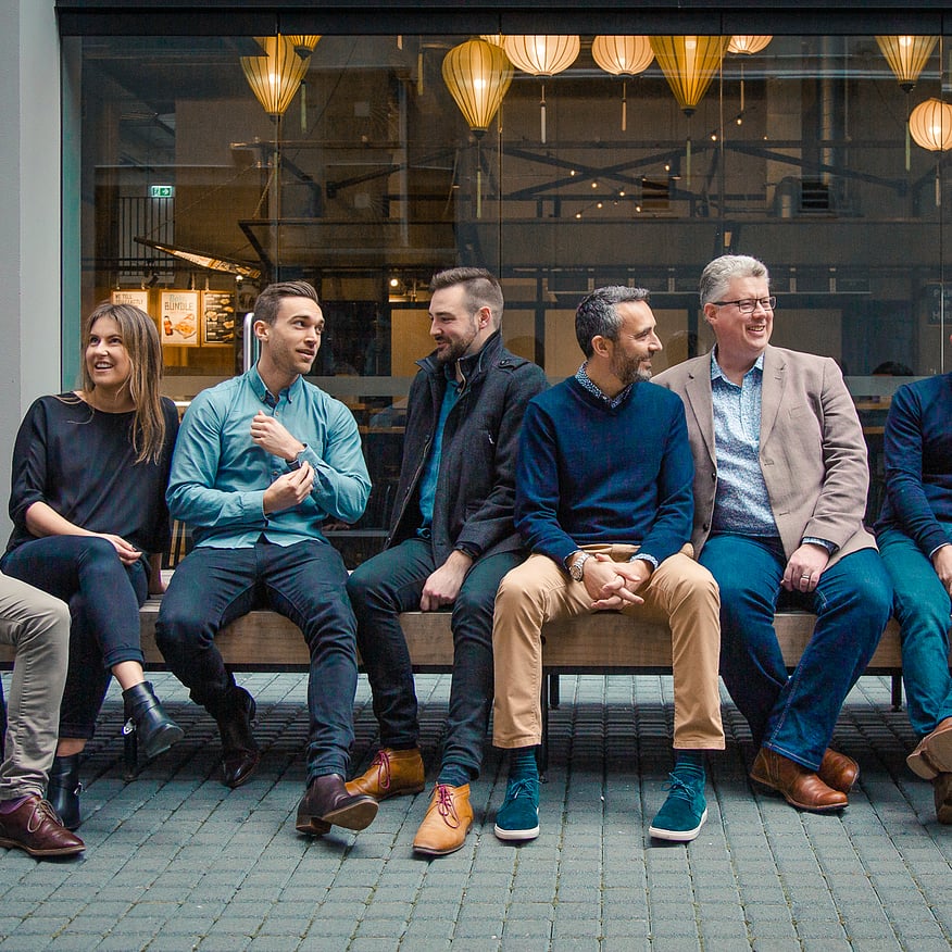 Group of nine casually dressed professionals sitting on a bench outside a modern caf&eacute;, engaged in conversation and laughter