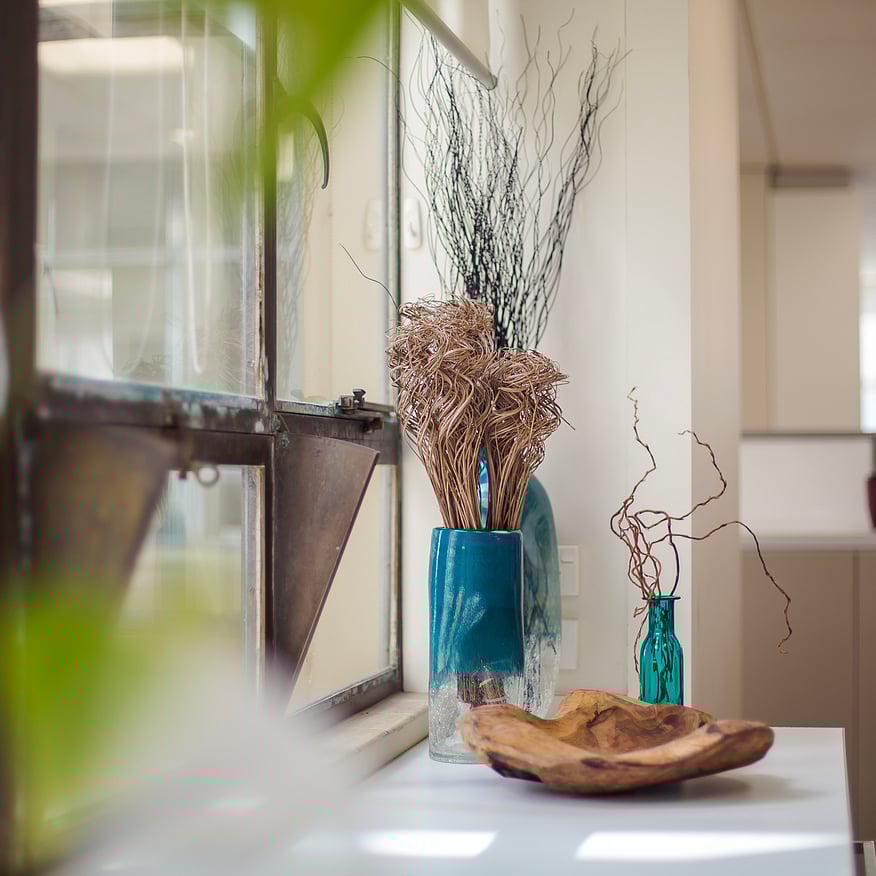 Bright office space with dried floral arrangements in blue glass vases and a wooden bowl on a white countertop near an open window