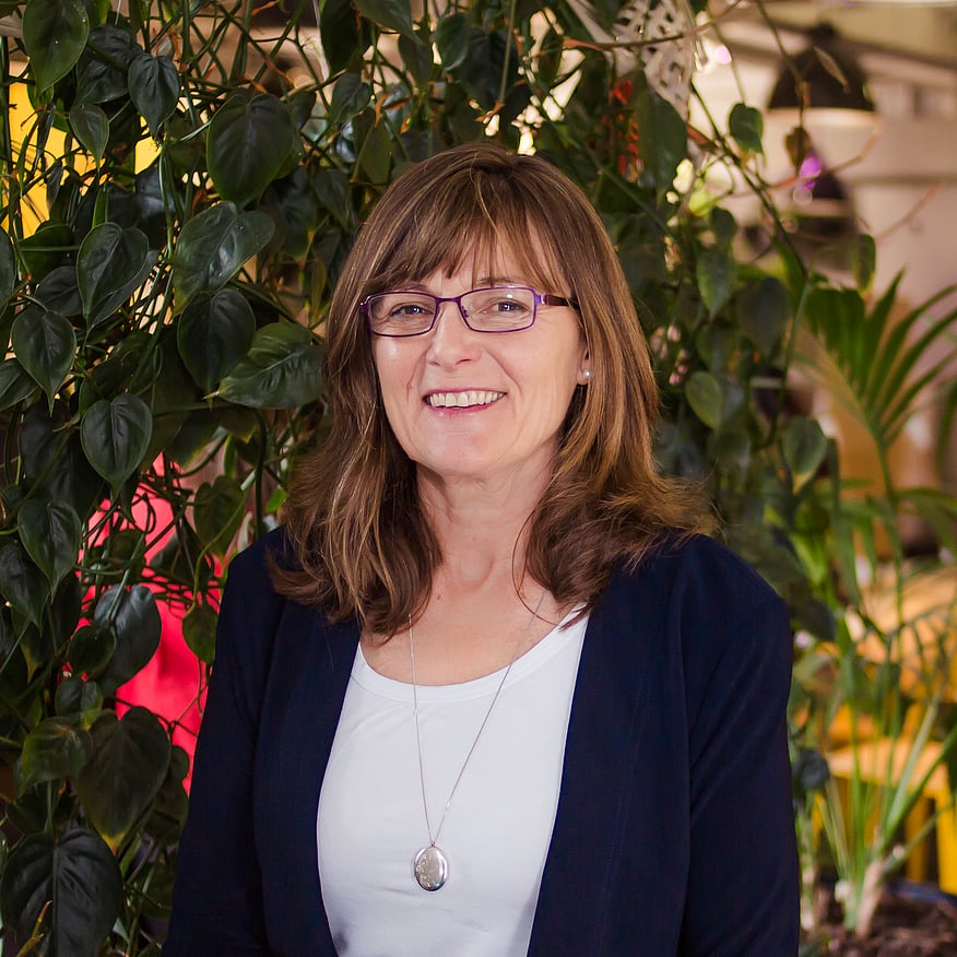Smiling woman with glasses and shoulder-length brown hair, wearing a navy blazer and white top, standing in front of green plants
