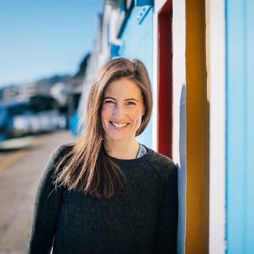 Smiling woman with long brown hair in a dark sweater leans against a colourful beach shed, with a marina and hillside buildings in the background