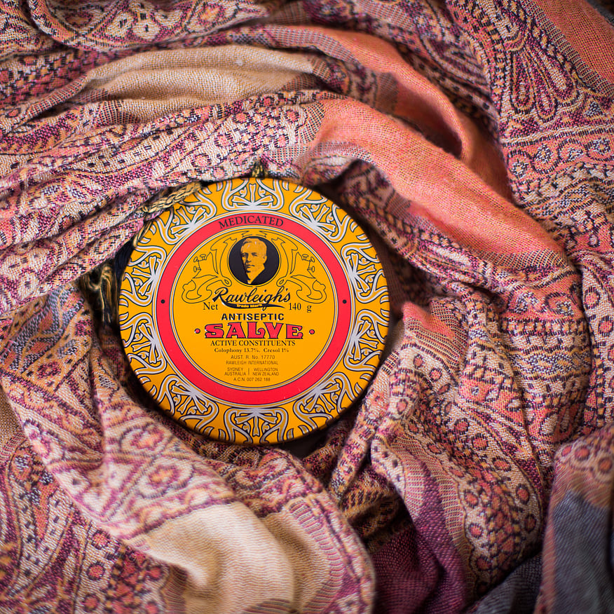 A tin of Rawleigh&rsquo;s Antiseptic Salve with a yellow and red vintage-style label sits nestled in a richly patterned fabric