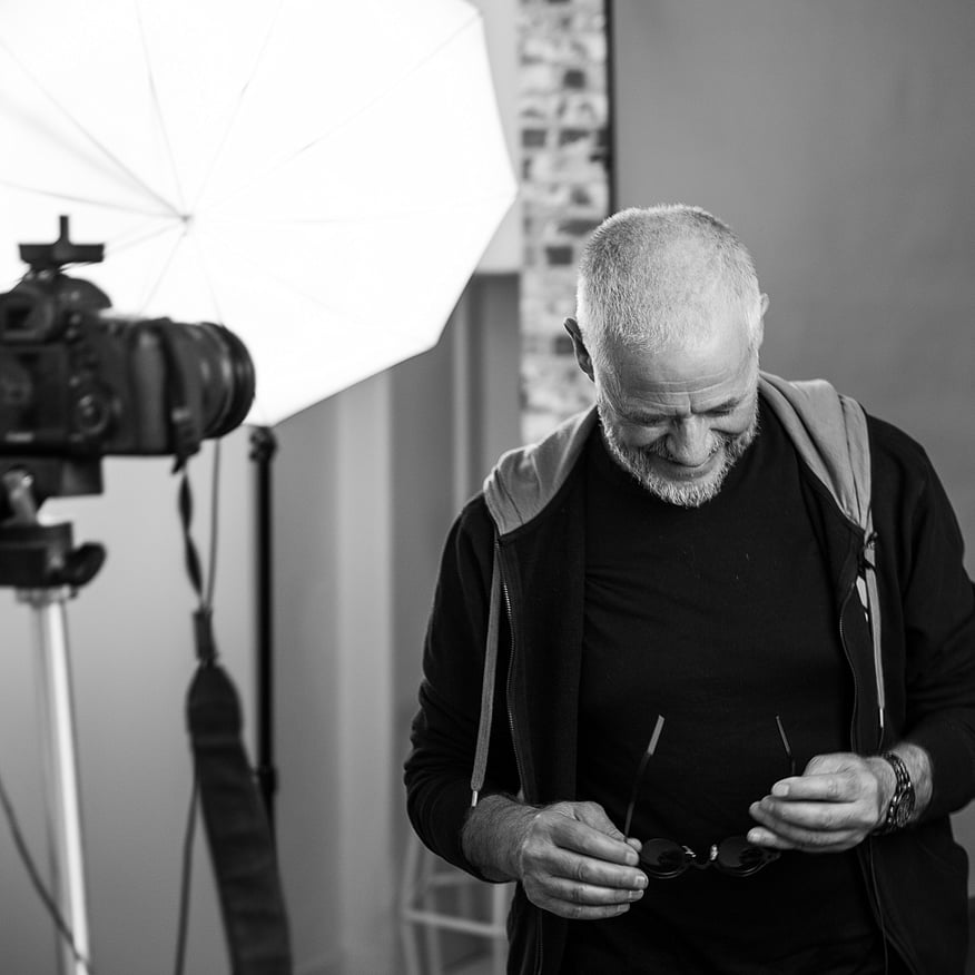 Black-and-white photo of a bearded man in a hoodie holding glasses in a photography studio with lighting and camera gear