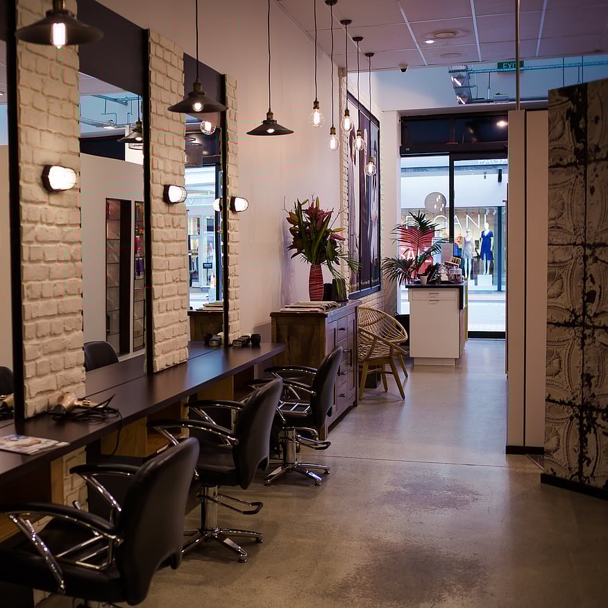 Modern hair salon with styling stations, large mirrors, black chairs, exposed brick walls, and pendant lighting, with a reception area in the background