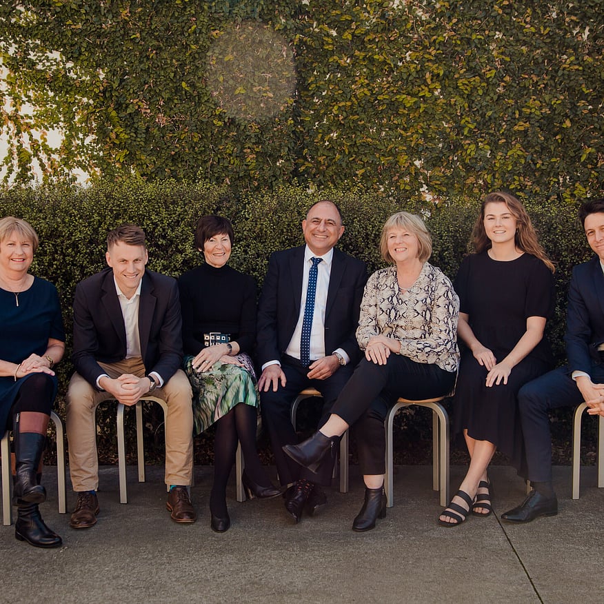 Seven professionally dressed people sitting on stools outdoors, smiling in front of a green hedge