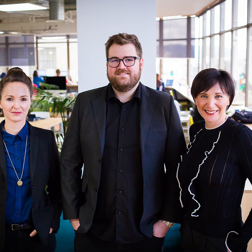 Three professionals, dressed in black suits, stand together in an office space with bright windows and plants in the background