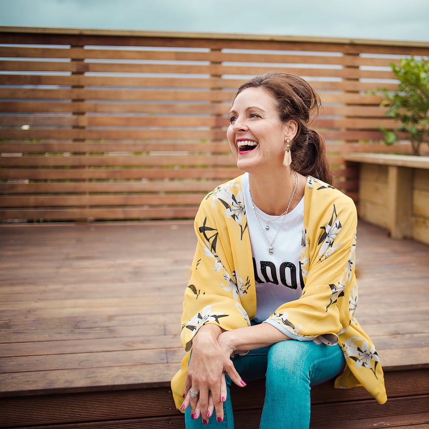 Woman sitting outdoors on a wooden deck, wearing a yellow floral kimono and a white t-shirt, smiling and laughing