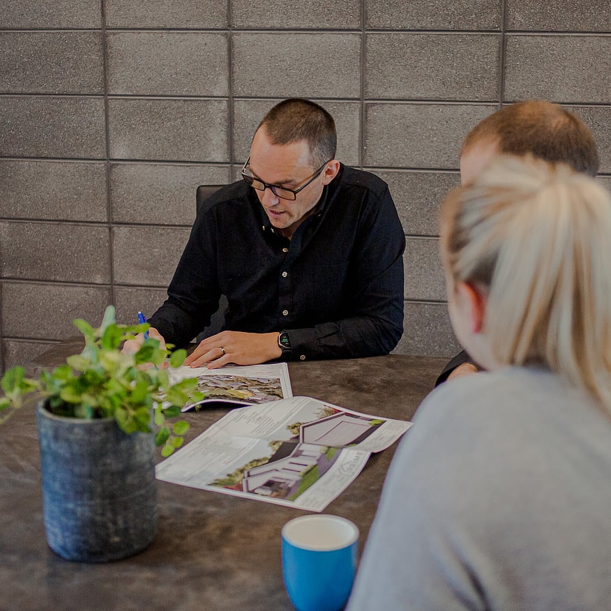 A group of three people in a meeting, reviewing architectural plans at a table with a plant and a blue cup visible