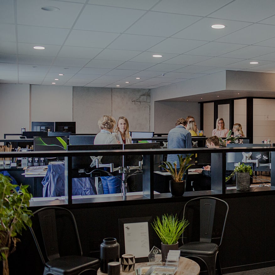 A modern office space with a group of people engaged in conversation, standing around a desk, while others work at their desks in the background