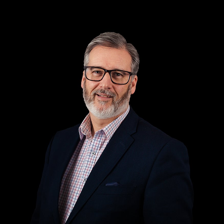 A man with glasses, a beard, and a checkered shirt under a dark blazer smiles confidently against a solid black background
