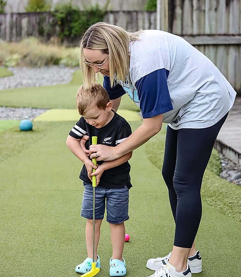 Mum and young son playing mini golf together, helping him hit the ball