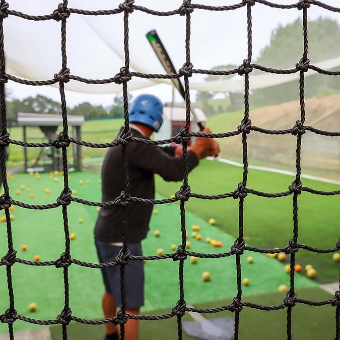 Man facing toward auto pitching machine in batting cage behind net