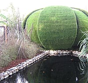 Kinetic grass-covered pod in closed position reflecting into black water pond