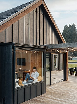This image shows the completed home with a deck, outdoor fairy lights on the pergola, a modern board-and-batten home, and the homeowner reading a book in the window seat.