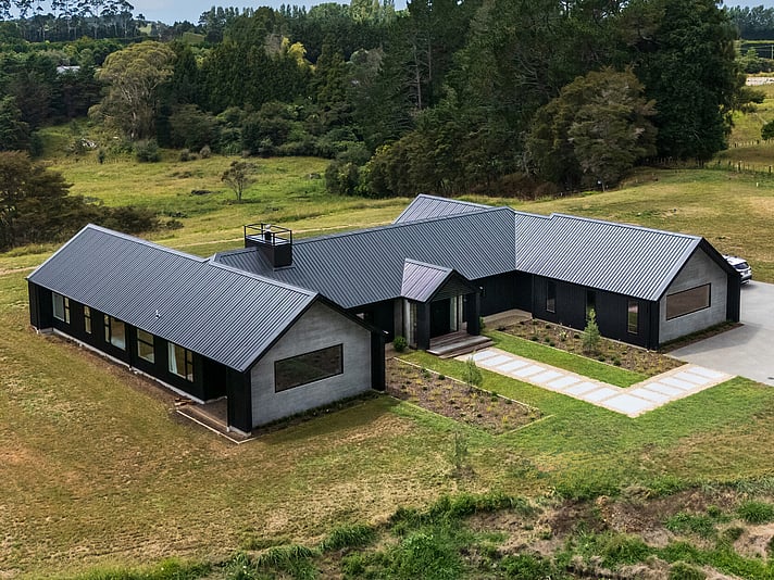 The project cover image shows a large new home built in rural Katikati. This new build shows a high level of workmanship completed by Straight Up Builders Tauranga. The image shows the exterior of the home, with wood-grain-effect concrete panels and black cladding, a new front door, and a Colorsteel roof.