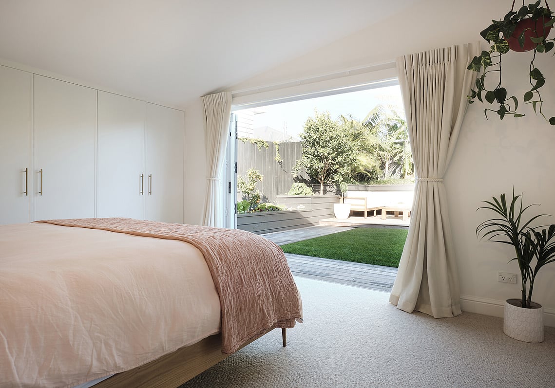 A newly renovated bedroom with new wardrobe doors, carpet, walls, curtains, bed, and bifold patio doors. The patio doors are open, looking out to the garden.