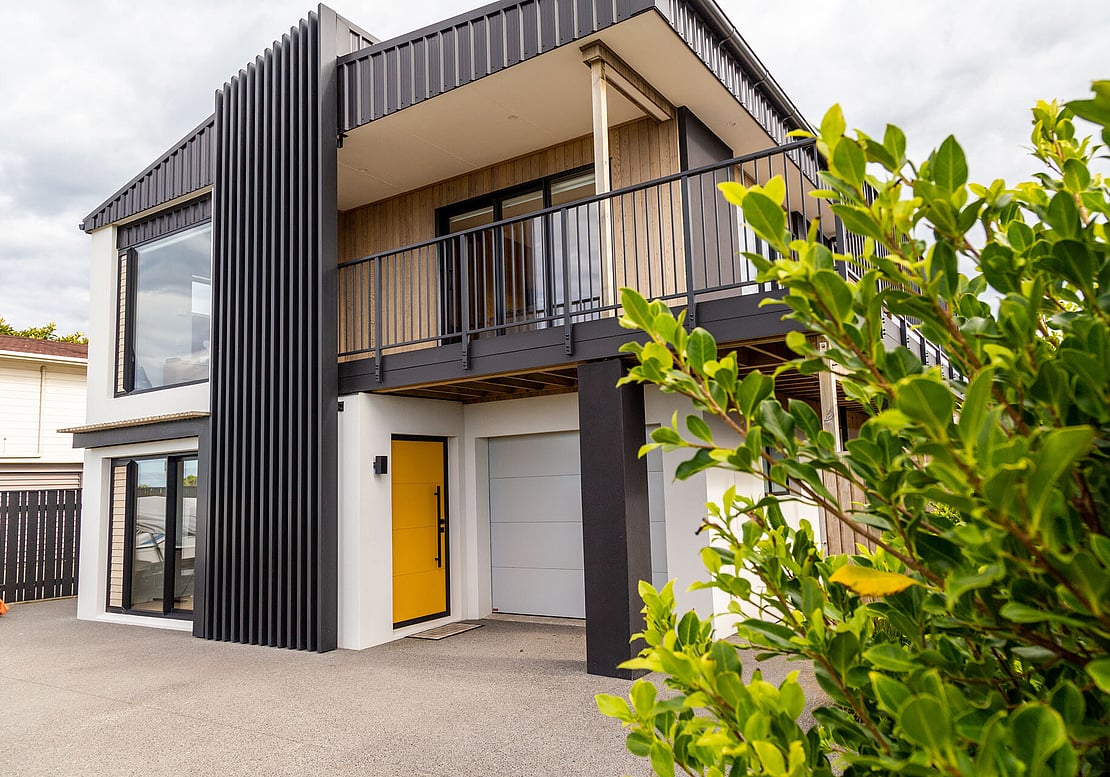 The image shows a recent renovation completed in Papamoa. It's a modern two-story house with a yellow front door, new cedar cladding and cream textured coating, and a deck on the second story.