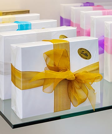 White boxes of Belgium chocolates placed on a glass shelf. Each box is wrapped with a different coloured ribbon.