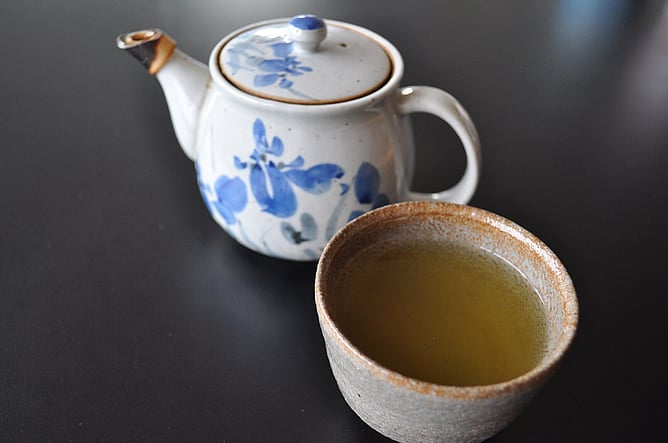 An earthware cup of green tea, with ceramic teapot behind