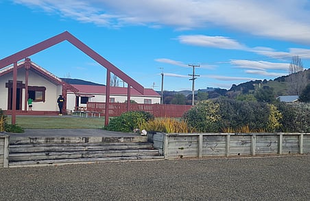 Marae with red carved entrance structure, buildings and landscaped grounds under blue sky with clouds