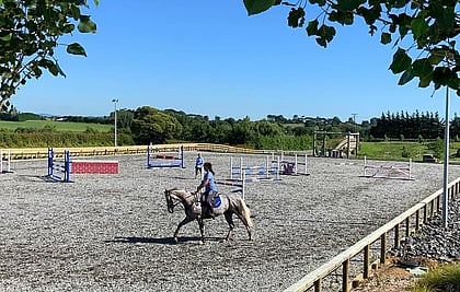 Lady rides a grey horse on an Ebb and flow equestrian arena on a summers day