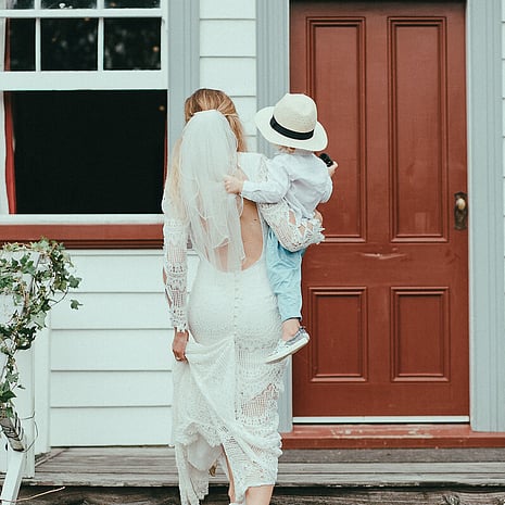 A bride walking up the steps towards the red wooden front door of the Kūaotunu Hall with a young child wearing a hat in her arms