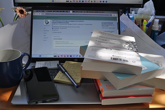 A laptop on a work desk piled with books and other work materials