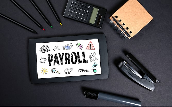 A flat lay view of pens, and other stationary including a tablet with Payroll written on the screen.